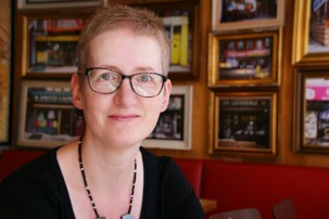 Dr Williams sitting in a room with painting covering the wall, wearing a black top, necklace and glasses.