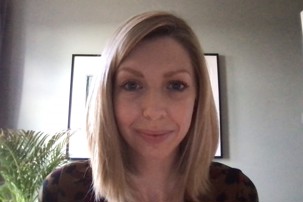 Dr Rice smiling to camera in front of a grey wall with a black and white artwork and a green plant is off to the side.